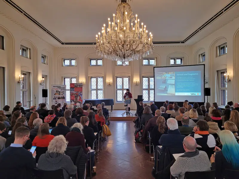 festlicher Saal mit vielen Personen, die auf Stühlen sitzen, rechts eine große Leinwand mit Projektion