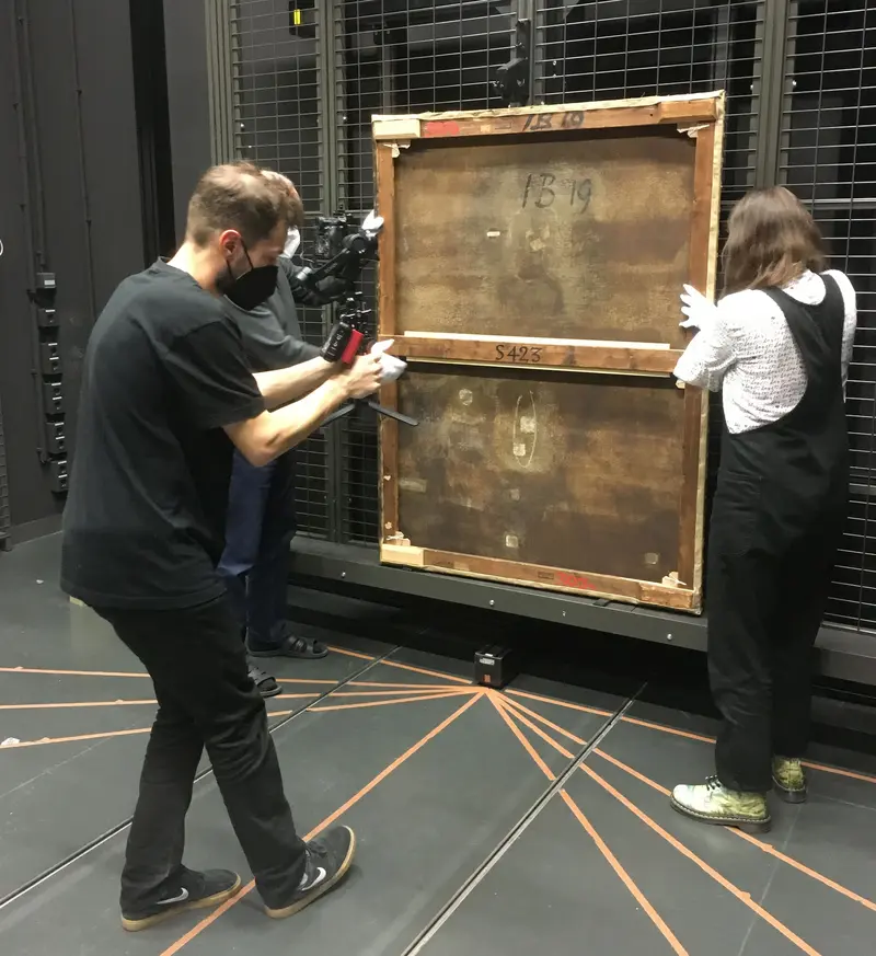 zwei Personen tragen ein Gemälde, von welchem man die Rückseite sieht